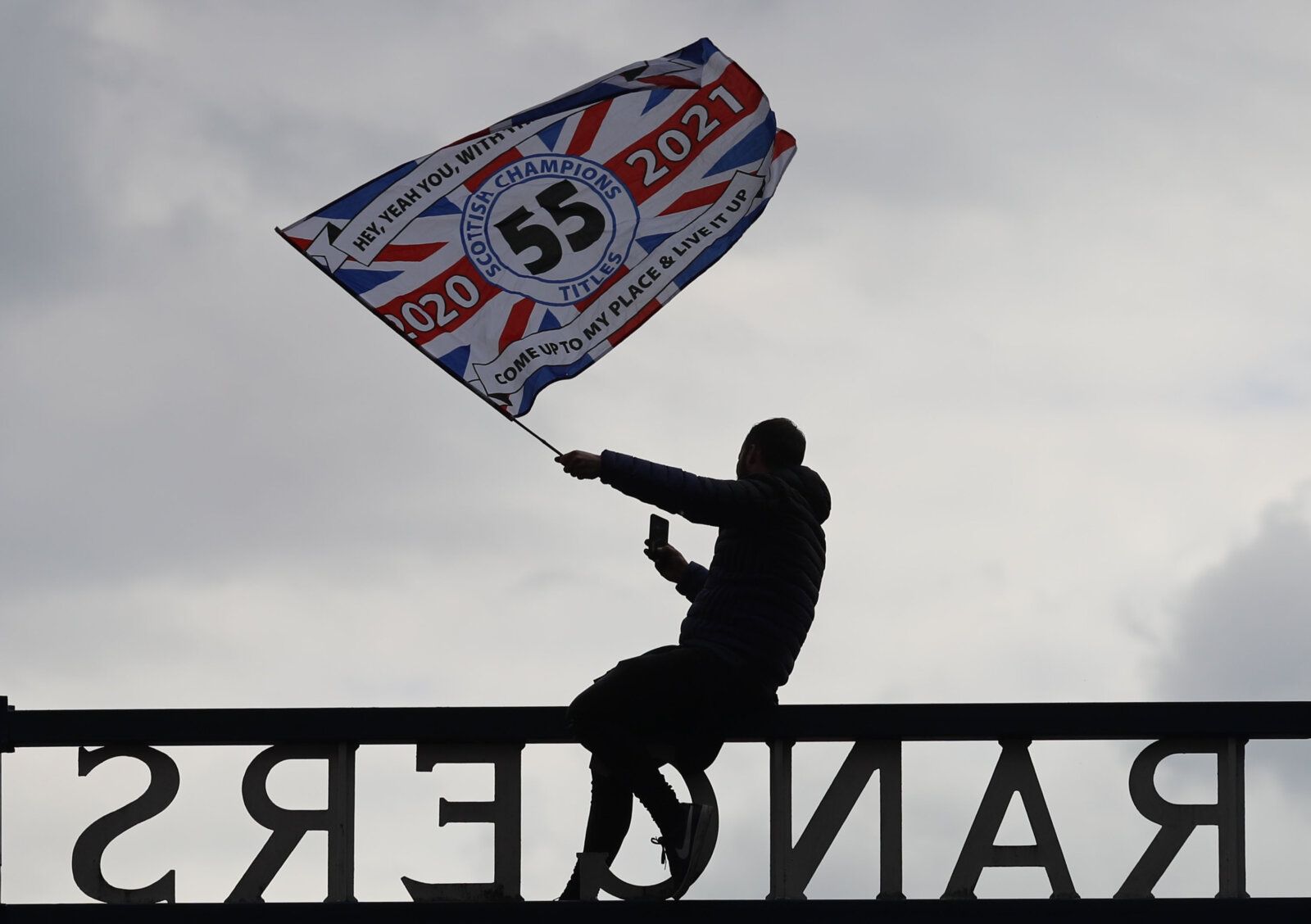 Soccer Football - Scottish Premiership - Rangers v Aberdeen - Ibrox, Glasgow, Scotland, Britain - May 15, 2021 A Rangers fan sat atop the stadium gate waves a flag as they react after winning the league REUTERS/Russell Cheyne