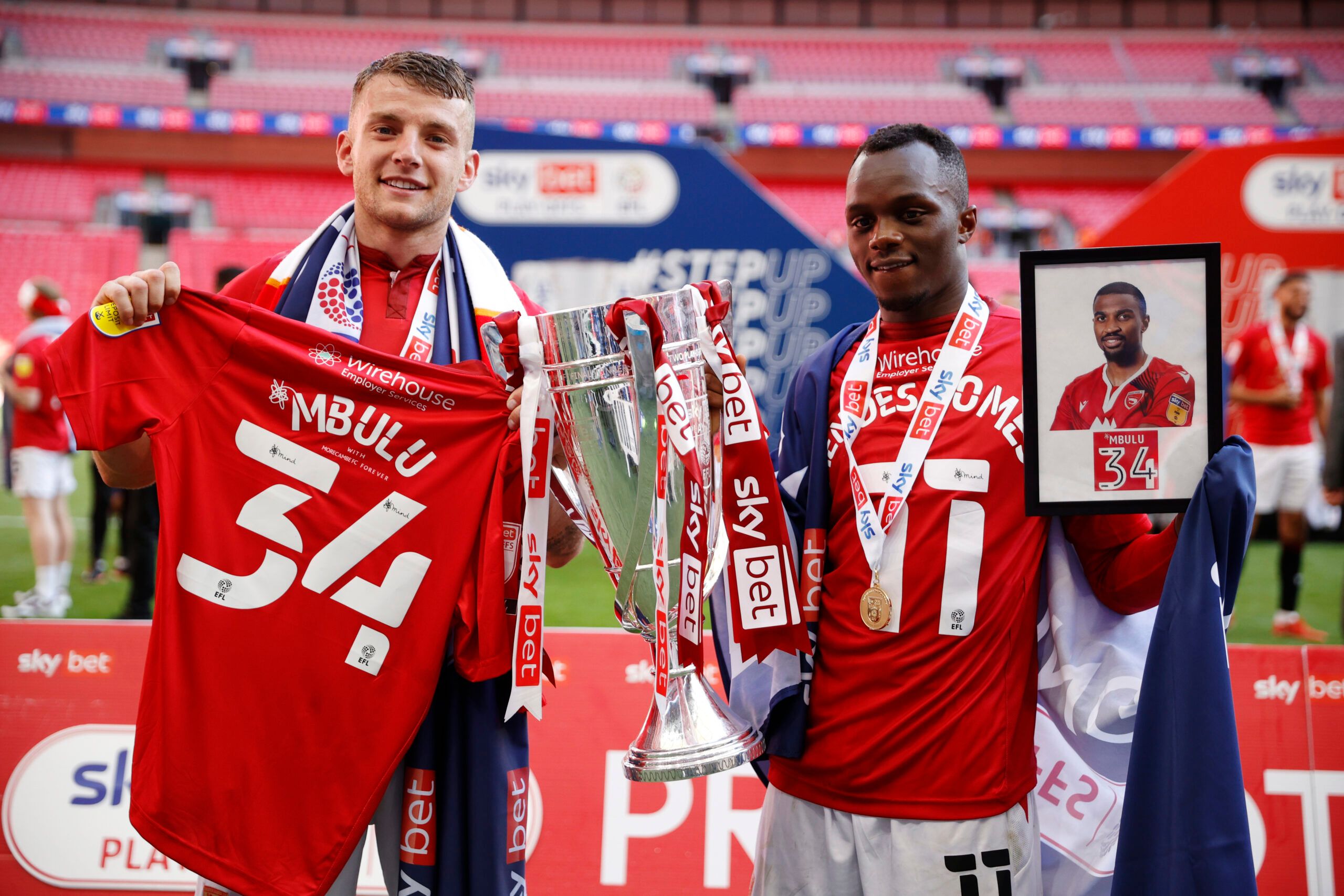 Soccer Football - League Two Play-Off Final - Morecambe v Newport County - Wembley Stadium, London, Britain - May 31, 2021 Morecambe's Carlos Mendes Gomes and Sam Lavelle pay tribute to former player Christian Mbulu's by holding up his shirt and photograph alongside the League Two Play-Off Final trophy Action Images/John Sibley EDITORIAL USE ONLY. No use with unauthorized audio, video, data, fixture lists, club/league logos or 'live' services. Online in-match use limited to 75 images, no video e